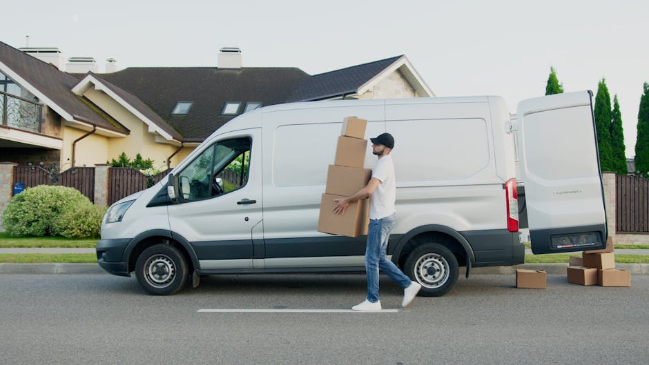 A man wearing casual clothing and a face mask is loading four cardboard boxes into the side of a white commercial van parked on a residential street. The van is positioned parallel to the curb with its side door open, revealing an interior prepared for home relocation. Three additional cardboard boxes are placed on the pavement near the vehicle, with some stacked inside and others on the ground. The background features modern two-story houses with pitched roofs, dormer windows, and a wooden fence, along with manicured bushes and tall, narrow trees. The scene is illuminated by natural daylight, capturing the loading process involved in furniture transport and packing during a house removal carried out by Removal Companies Uxbridge, as part of their same-day removals service in Uxbridge.