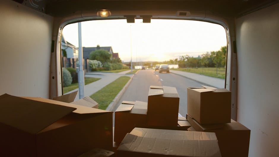 A man wearing casual clothing and a face mask is loading four cardboard boxes into the side of a white commercial van parked on a residential street. The van is positioned parallel to the curb with its side door open, revealing an interior prepared for home relocation. Three additional cardboard boxes are placed on the pavement near the vehicle, with some stacked inside and others on the ground. The background features modern two-story houses with pitched roofs, dormer windows, and a wooden fence, along with manicured bushes and tall, narrow trees. The scene is illuminated by natural daylight, capturing the loading process involved in furniture transport and packing during a house removal carried out by Removal Companies Uxbridge, as part of their same-day removals service in Uxbridge.