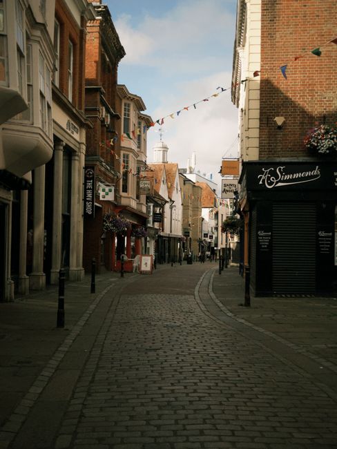 A narrow cobbled street in a town centre, lined with multi-storey brick and stucco buildings featuring shopfronts and hanging flower baskets. The street is decorated with small triangular flags strung across the sky, which is partly cloudy with sunlight casting shadows on the pavement. On the right side, there is a closed black roller shutter with the name 'H. Simmonds' displayed above it, while on the left, various commercial signs are visible. The street appears pedestrianised and is part of a typical high street, suitable for home relocation and furniture transport activities. The scene is captured during daytime with ambient lighting indicating daytime hours. This environment is typical for house removals, packing, and moving logistics, as shown in the context of a professional removal services website for Uxbridge.