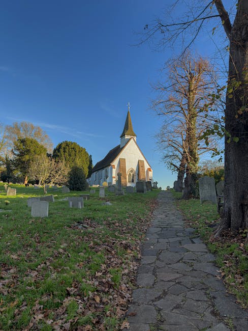 A narrow, uneven stone pathway leading uphill through a churchyard with grass and fallen leaves on either side. To the right of the path, there are several leafless trees and a few small headstones. To the left, there are additional gravestones and some dense, green foliage. At the top of the path stands a white church with a tall, pointed steeple, surrounded by well-maintained grass and distant trees under a clear blue sky. The scene is illuminated by natural daylight, capturing the peaceful atmosphere of a historic rural church setting. This image on the Uxbridge house removals guide for UB8 High Street moves highlights the typical environment associated with home relocation or furniture transport within a traditional village context, as managed by Removal Companies Uxbridge.